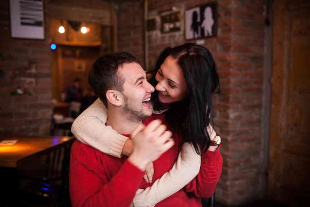 Pareja sonriente en un café, ella abraza a él por la espalda mientras ambos ríen