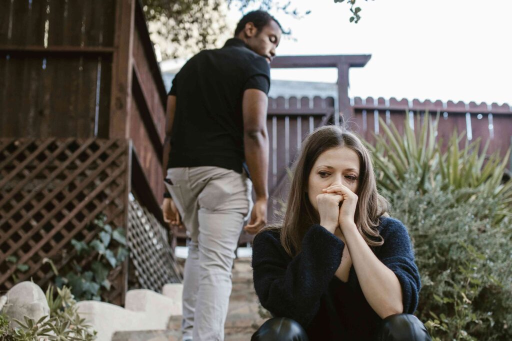 Mujer angustiada sentada con las manos en la boca mientras su pareja se aleja caminando por un jardín